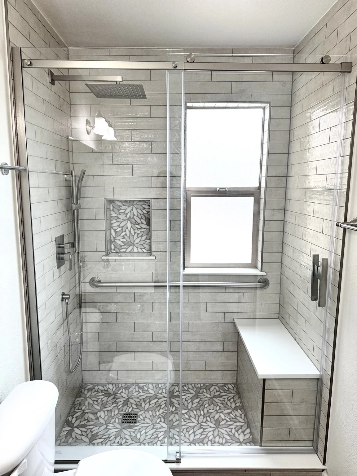 Completed custom shower with gray tile, window, glass enclosure, and built-in bench